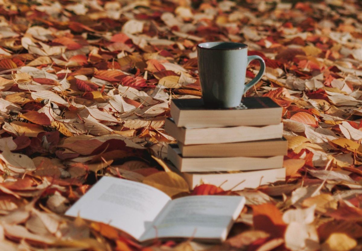 A cup of coffee placed on top of a pile of books. The books are laying on a bed of fall leaves.
