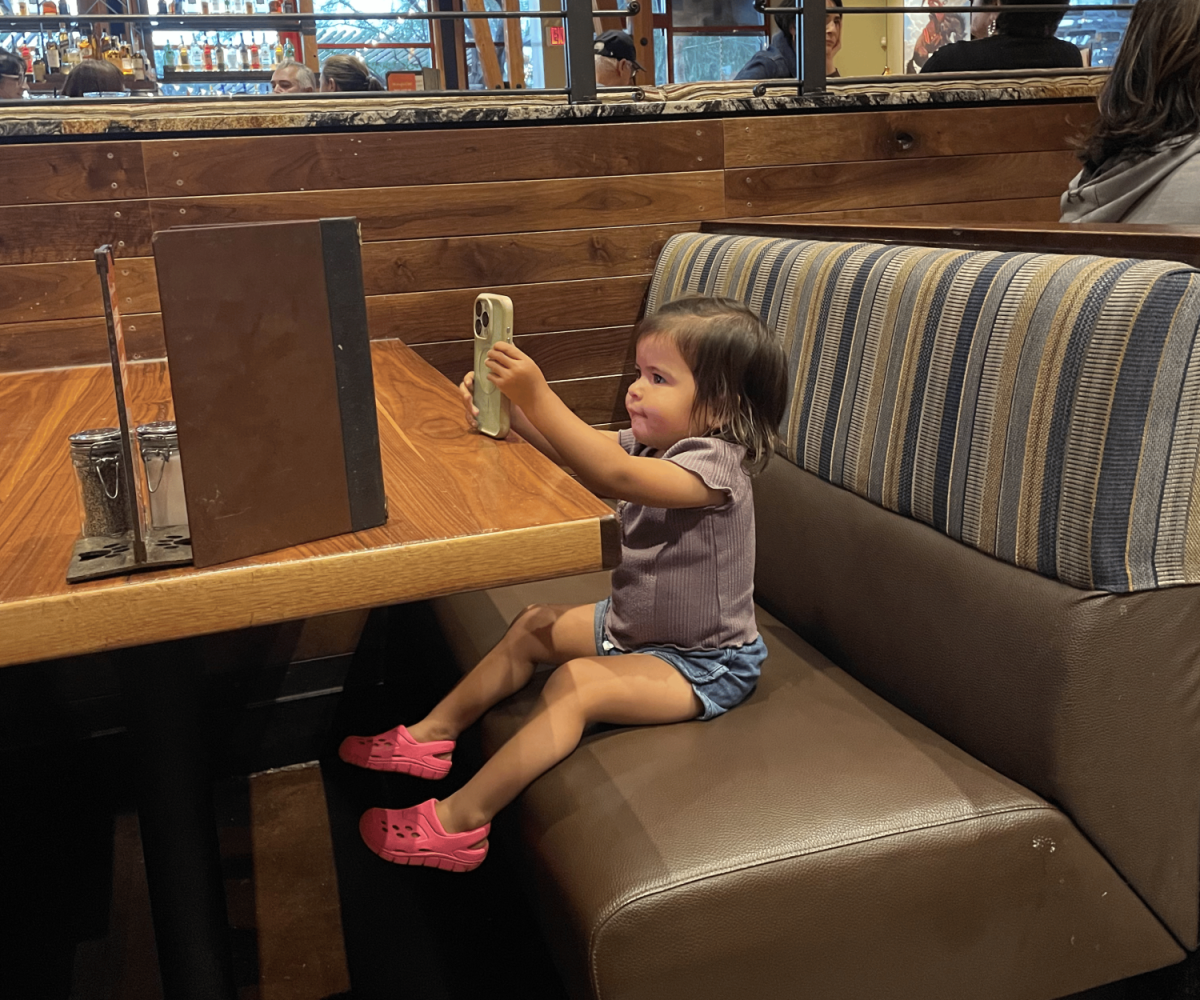 A toddler focuses on a phone screen while dining out, highlighting how digital devices have become a part of everyday life.