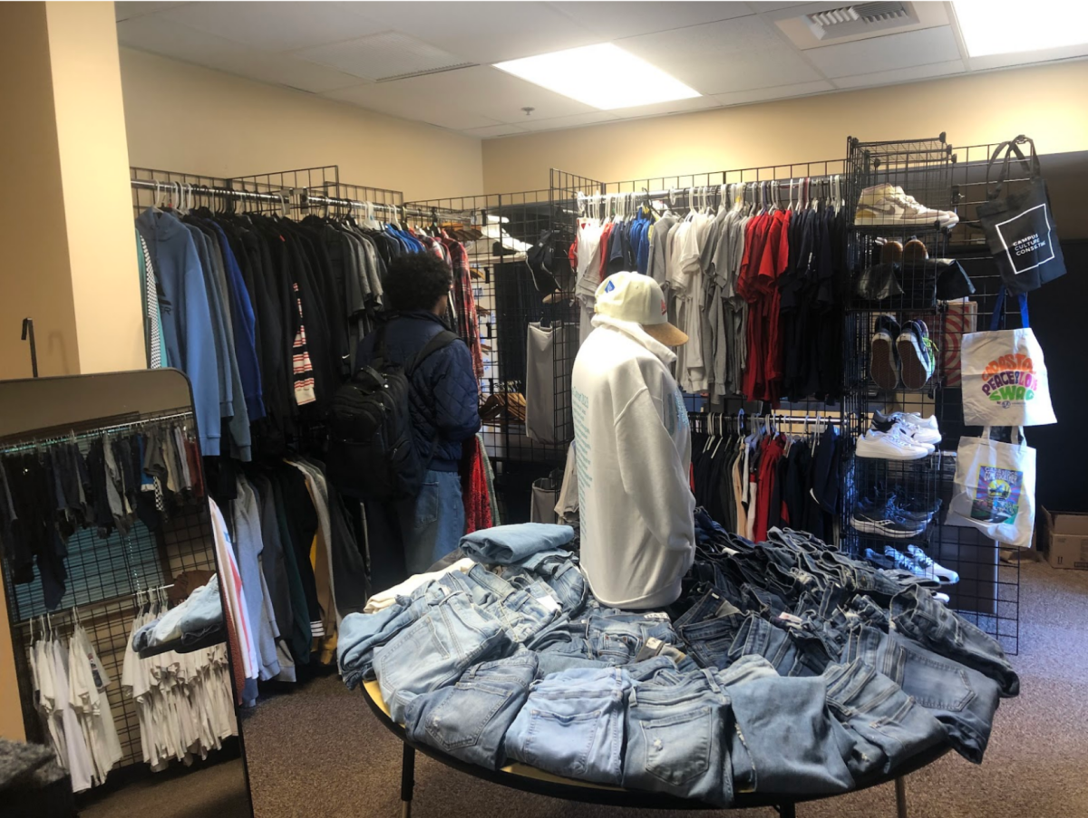A student browses the racks of clothing at Mustang Locker, with a central table overflowing with jeans of different sizes.